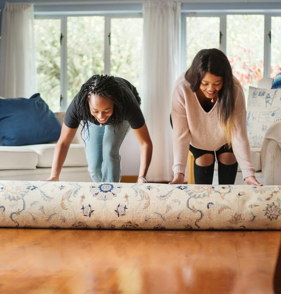 Two women unrolling area rug onto wood floor